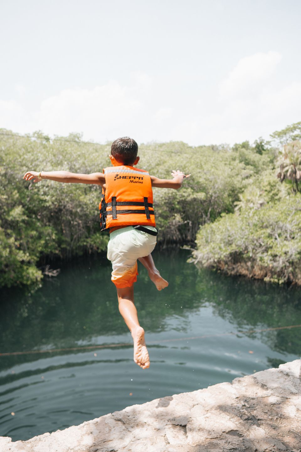 A child in an orange life vest jumping from a ledge into a green cenote, arms out, from behind
