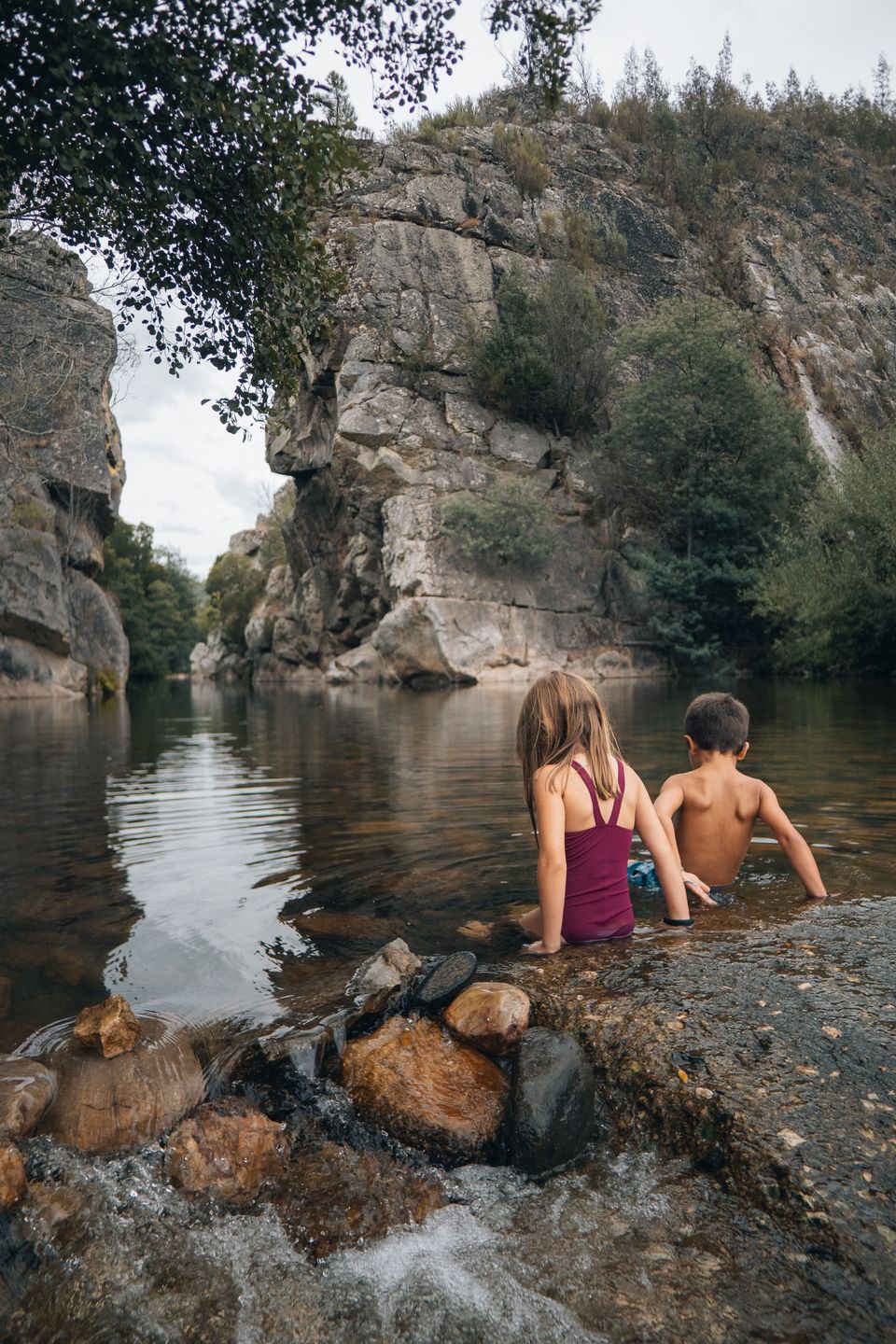 Two kids sitting on rocks at the edge of a river gorge, from behind